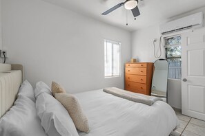 Ein Schlafzimmer mit einem Bett, einer Kommode, einem Fenster mit Jalousien, einer Klimaanlage und einem Deckenventilator.