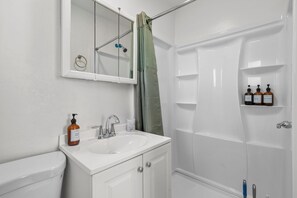 Ein Badezimmer mit einem weißen Waschbecken, einem Spiegel, einer Toilette, einer Dusche mit Glastür und einem Ablagebereich mit drei Toilettenartikeln.