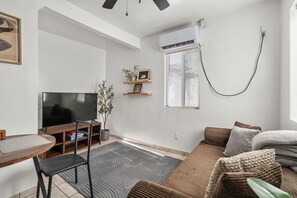 Ein modernes Wohnzimmer mit Flachbildfernseher, Essbereich mit Tisch und Stühlen, einer Couch mit Kissen, einer Topfpflanze und einem Fenster mit Jalousien.