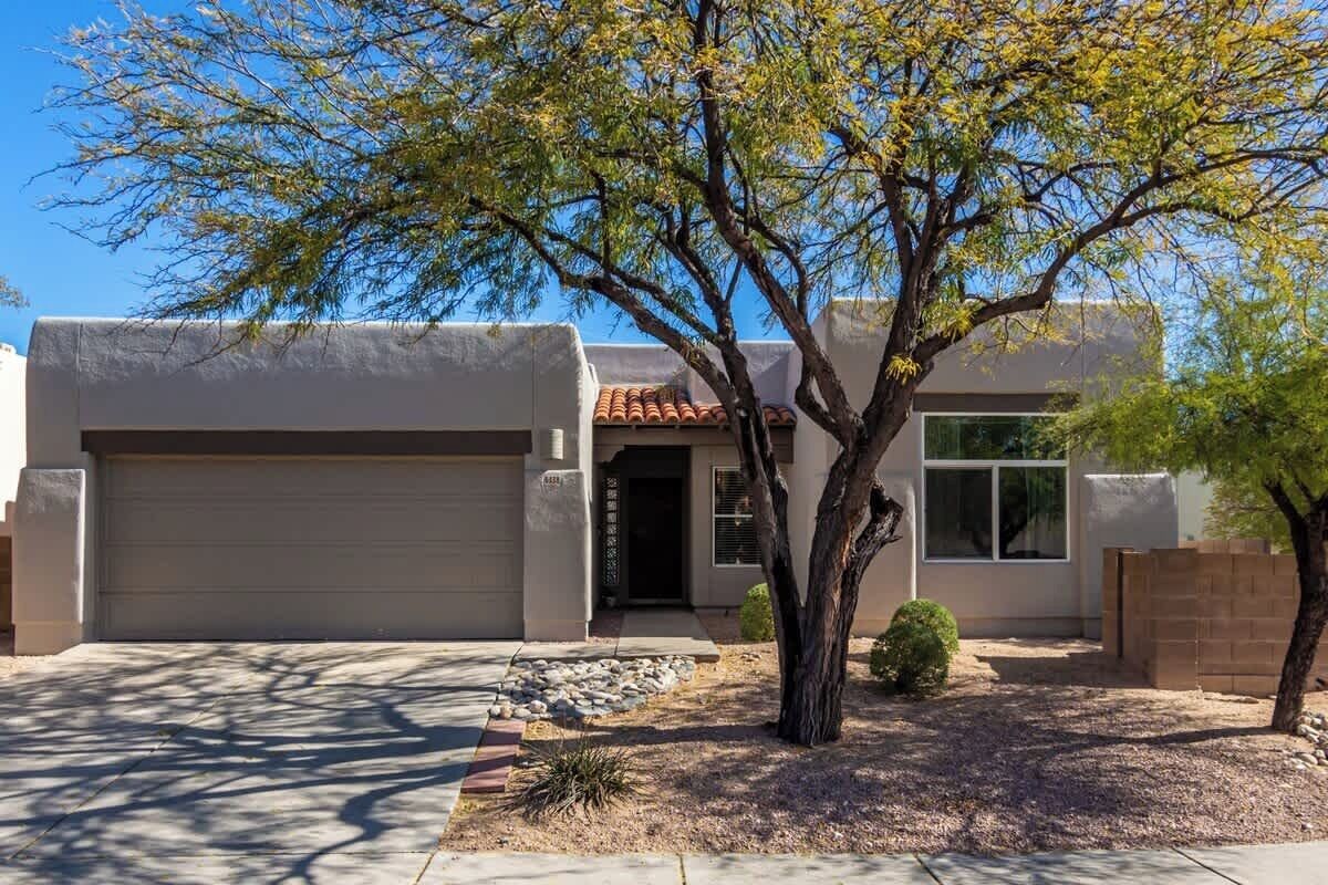 Une maison moderne à deux étages, dotée d’un garage, d’un petit jardin devant avec un arbre, et surplombée d’un ciel bleu dégagé.
