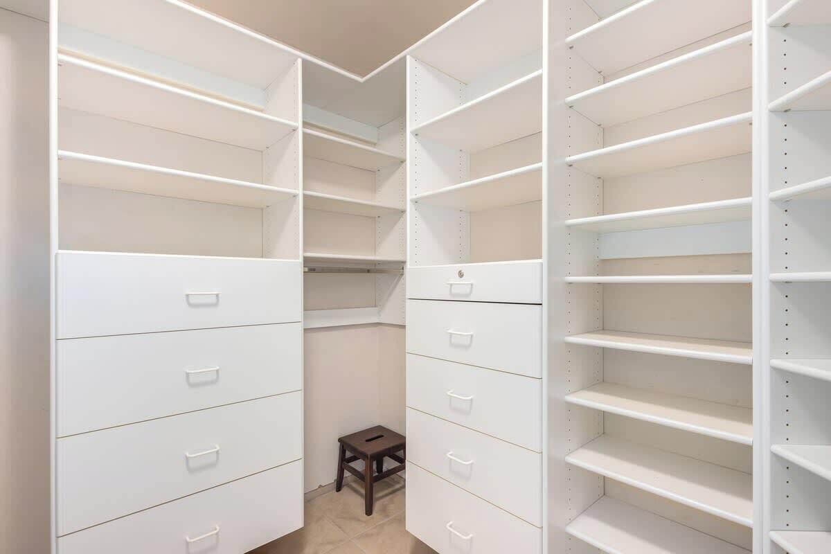 Ein begehbarer Kleiderschrank mit mehreren Regalen und Schubladen, einem kleinen Hocker und einer Tür zu einem anderen Raum.