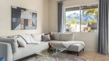 Ein modernes Wohnzimmer mit einem Ecksofa, einem Glastisch, einem Fellteppich und einem großen Fenster mit Blick auf die Berge.