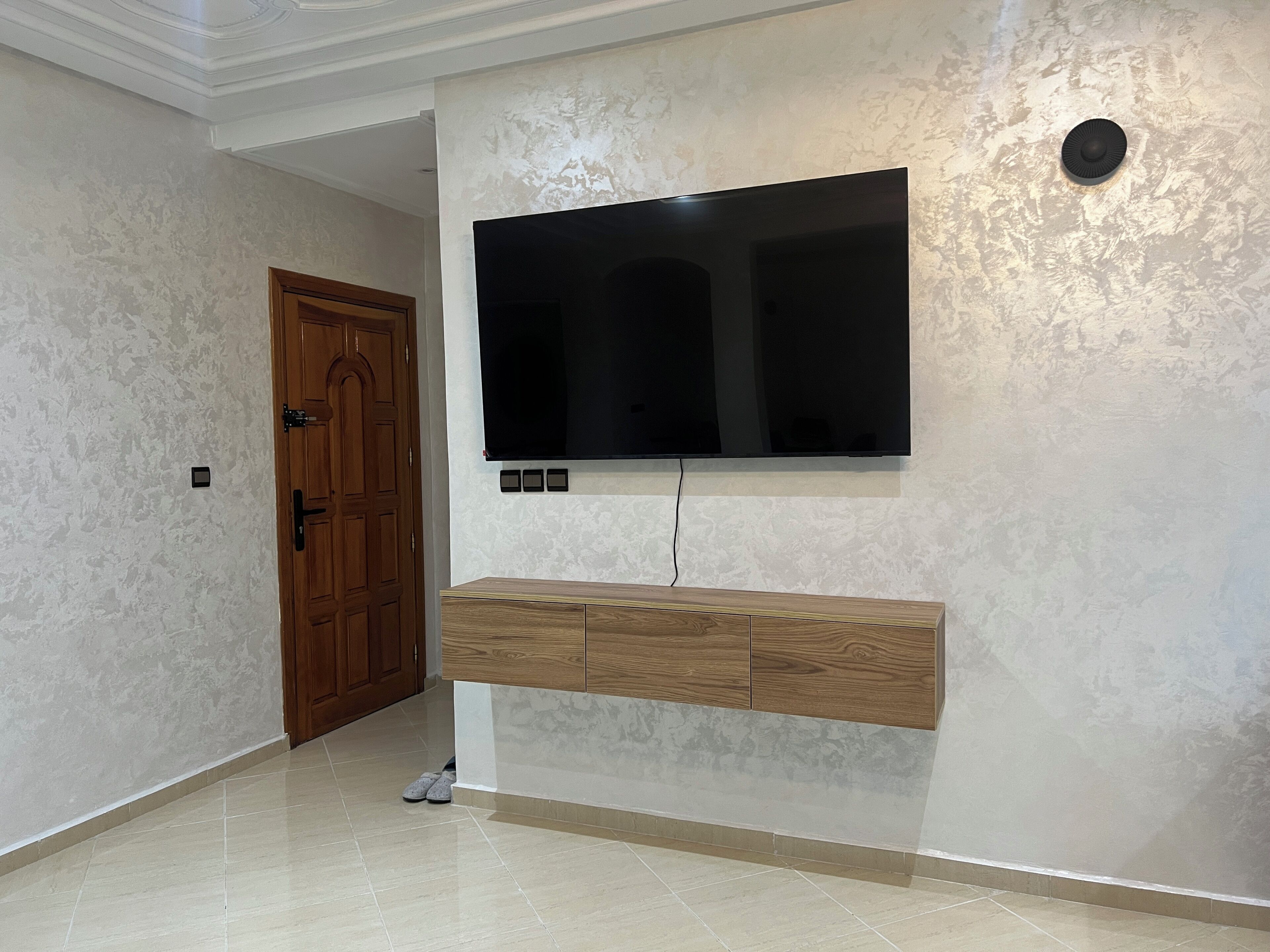 A modern living room with a flat-screen TV mounted on the wall, wooden cabinets below, and a tiled floor.