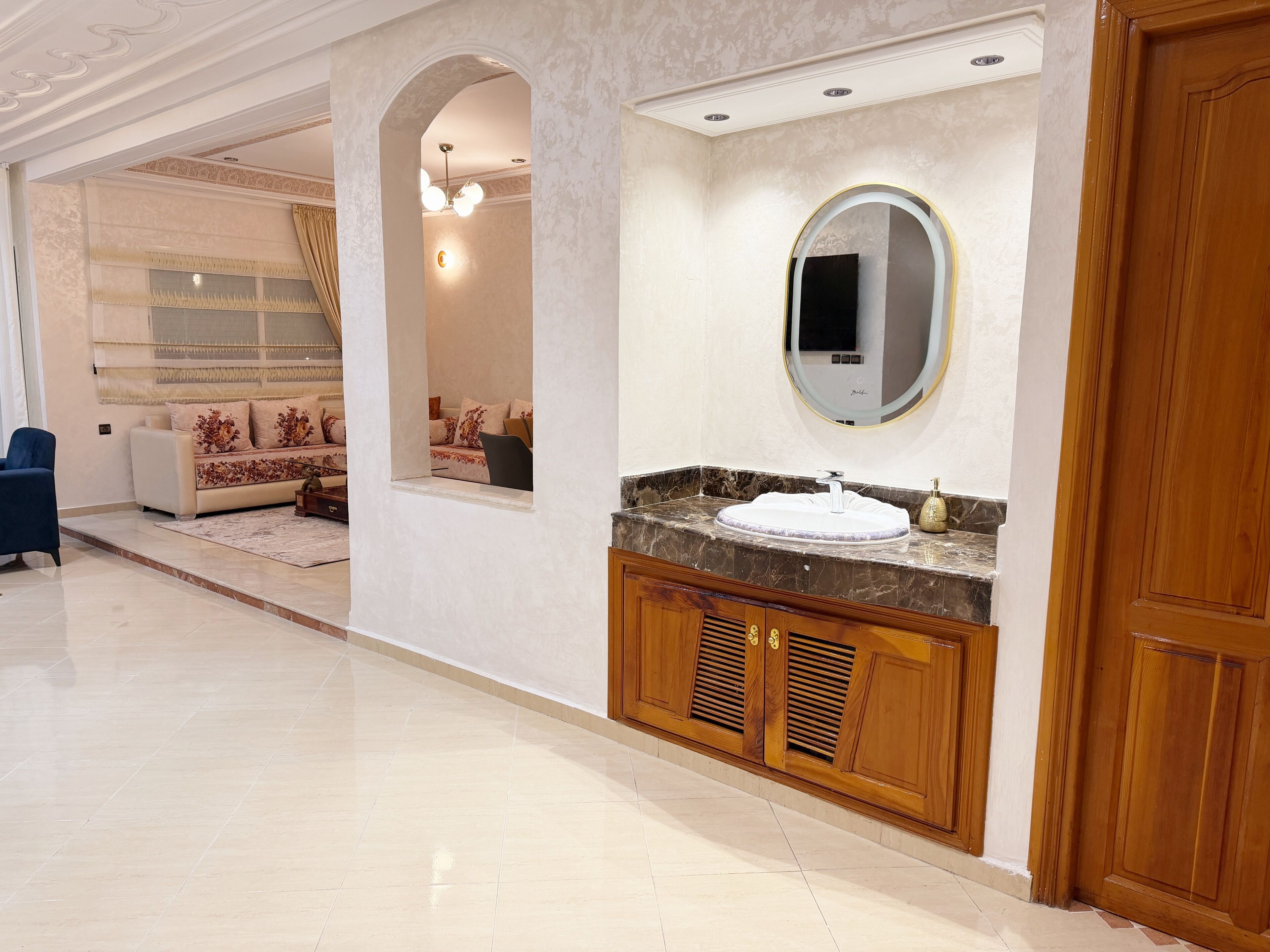 A spacious hotel room with a marble floor, a large round mirror, a wooden vanity with a sink, and a sofa in the background.