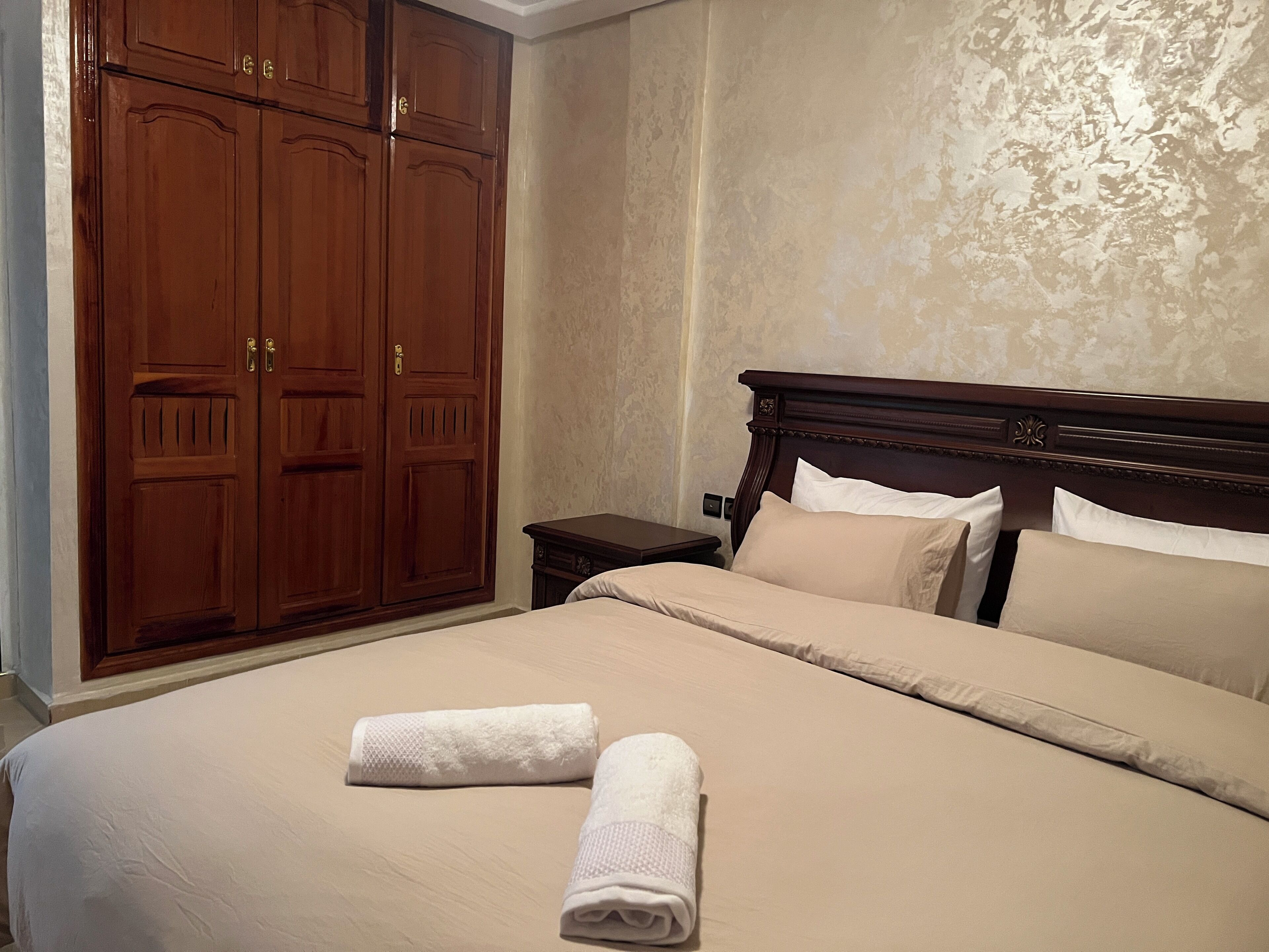 A bedroom with a large wooden wardrobe, a bed with beige bedding, and a nightstand.