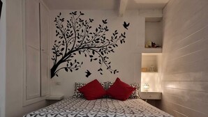 Un dormitorio con cama, almohadas rojas, un mural en la pared de un árbol con pájaros y una mesita de noche con lámpara.