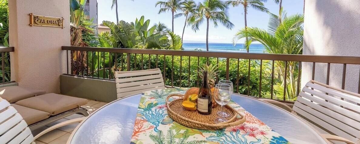 Un balcon avec une table dressée pour deux personnes, agrémentée d’une bouteille et d’un verre, et offrant une vue tropicale sur des palmiers et l’océan.