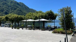Un área de estar al aire libre con mesas y sombrillas, rodeada de plantas en macetas y árboles, con vista a un lago y montañas al fondo.