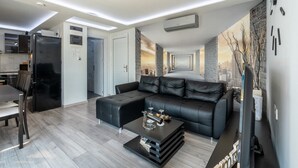 Een moderne woonkamer met een zwarte leren bank, een eettafel met stoelen, een bijzettafeltje en een muurschildering van een stadsgezicht.