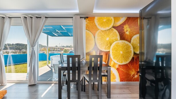 Een eetgedeelte met een tafel en stoelen, uitzicht door grote ramen, en een wand met een fruitmotief.