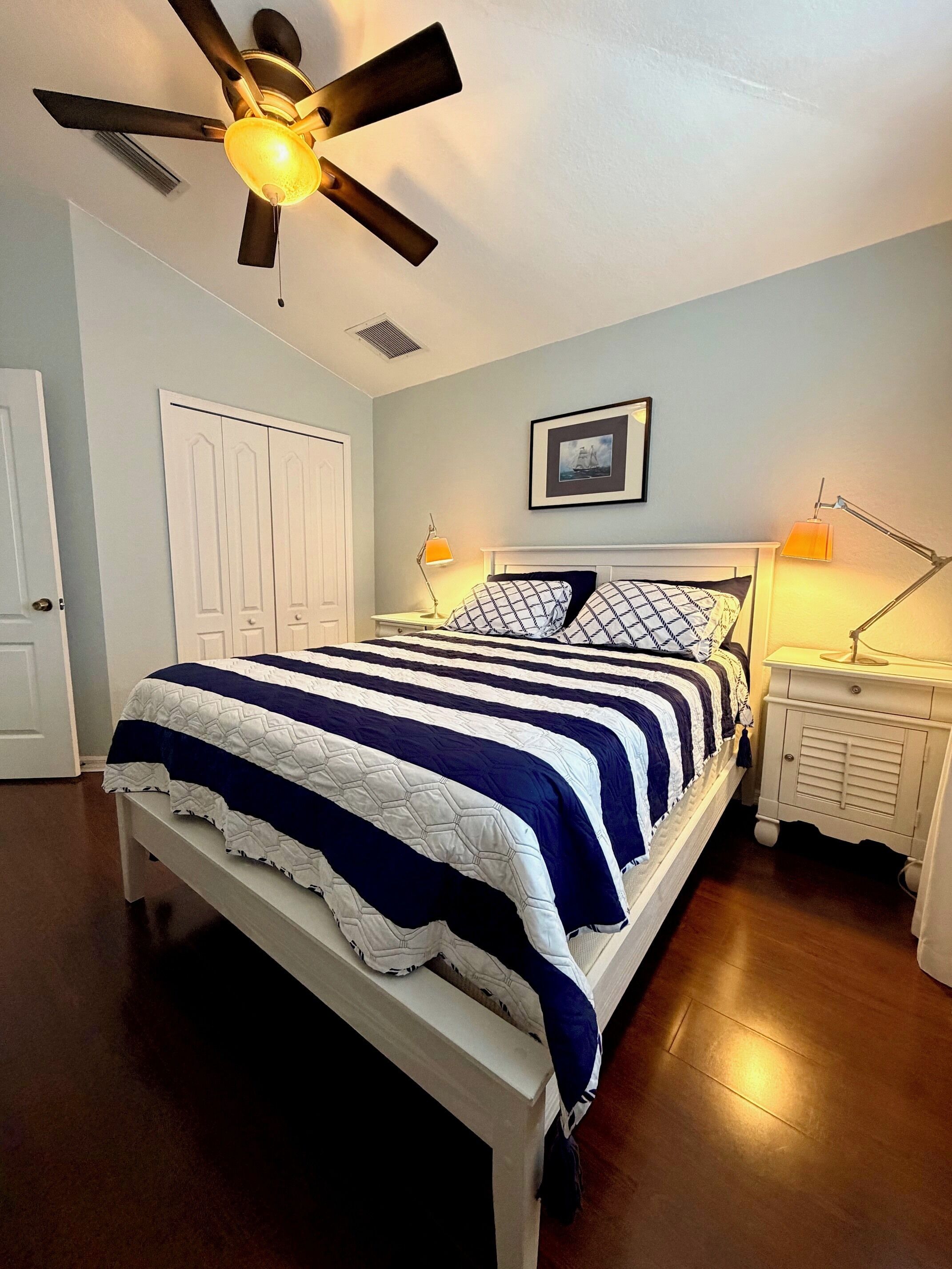 Un dormitorio con cama, ventilador de techo, mesita de noche con lámpara y una puerta.