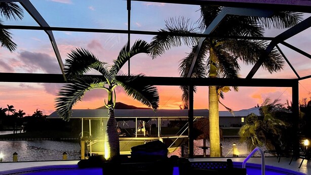 Ein Poolbereich mit Liegestühlen, einer Glasveranda und Blick auf einen Sonnenuntergang über dem Wasser.