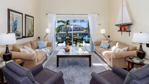 Ein geräumiges Wohnzimmer mit zwei Sofas, einem Couchtisch und Blick auf einen Pool sowie Palmen durch große Glasfenster.
