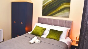 Una cama bien hecha con colcha gris, dos almohadas verdes y toallas enrolladas. Un armario azul grande está a la izquierda, y un cuadro cuelga sobre la cama.