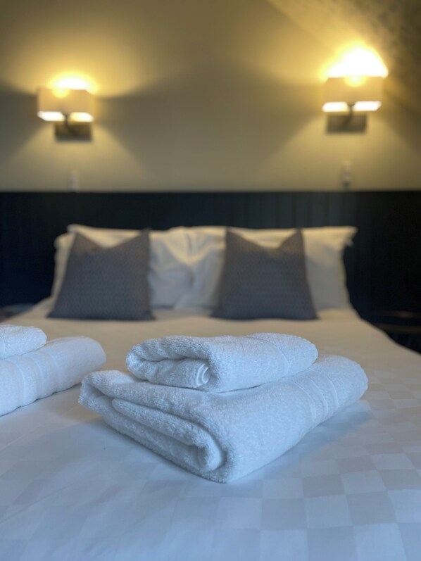 A neatly made bed with white linens and folded towels, two wall-mounted lamps, and a dark headboard.