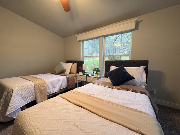 A bedroom with two beds, a nightstand, a lamp, and a window with a view of trees.