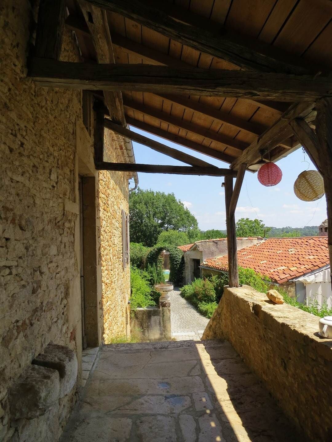 A stone pathway leading to a rustic building with a wooden roof and a view of a red-tiled roofed area in the distance.