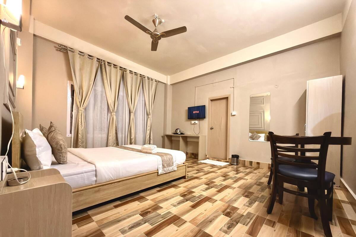 A hotel room with a bed, a desk, a chair, a ceiling fan, a TV, and a wooden parquet floor.
