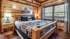 A wooden cabin bedroom with a bed, two bedside tables, a window with a view, and a ceiling with exposed beams. - Incredible Family Cabin-Gamers Lounge & Theater Rm (Sevierville)