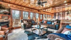 A spacious living room with wooden ceilings, large windows, a stone fireplace, leather sofas, and a central coffee table. - Incredible Family Cabin-Gamers Lounge & Theater Rm (Sevierville)