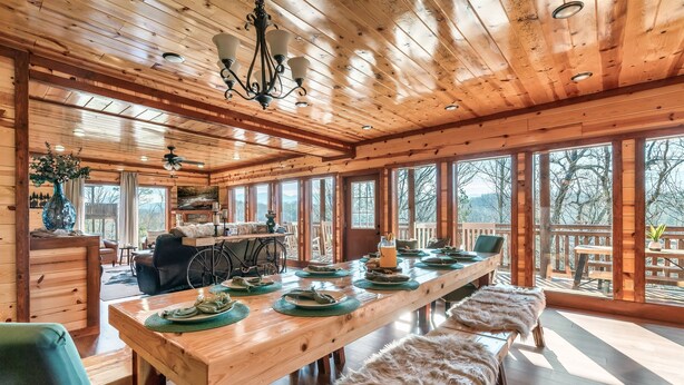 A spacious wooden dining area with a long table set for a meal, large windows offering a view of the outdoors, and a cozy seating arrangement with benches. - Incredible Family Cabin-Gamers Lounge & Theater Rm (Sevierville)