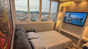 Ein Zimmer mit einem Bett, einem Fernseher, einem Fenster mit Blick auf die Stadt und einem Gemälde an der Wand.