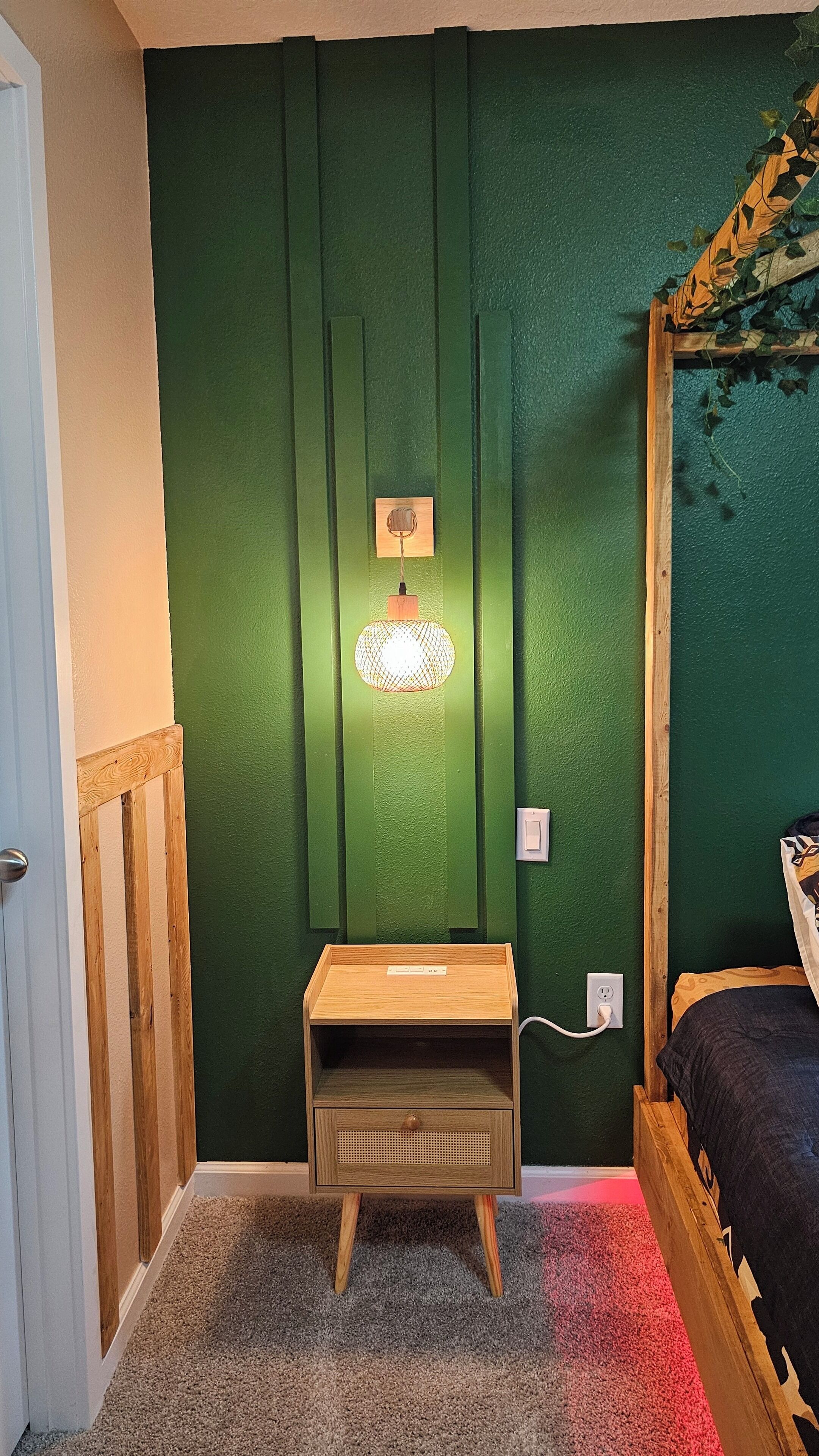 A bedroom with a green wall, a wooden nightstand, a bed with a patterned bedspread, and a wall-mounted lamp.