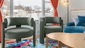 Ein Zimmer mit zwei grünen Sesseln, einem blauen Sofa, einem Holztisch und einem farbenfrohen Teppich.