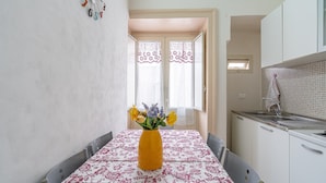 A dining area with a table set for two, a vase with flowers, and a kitchen in the background.