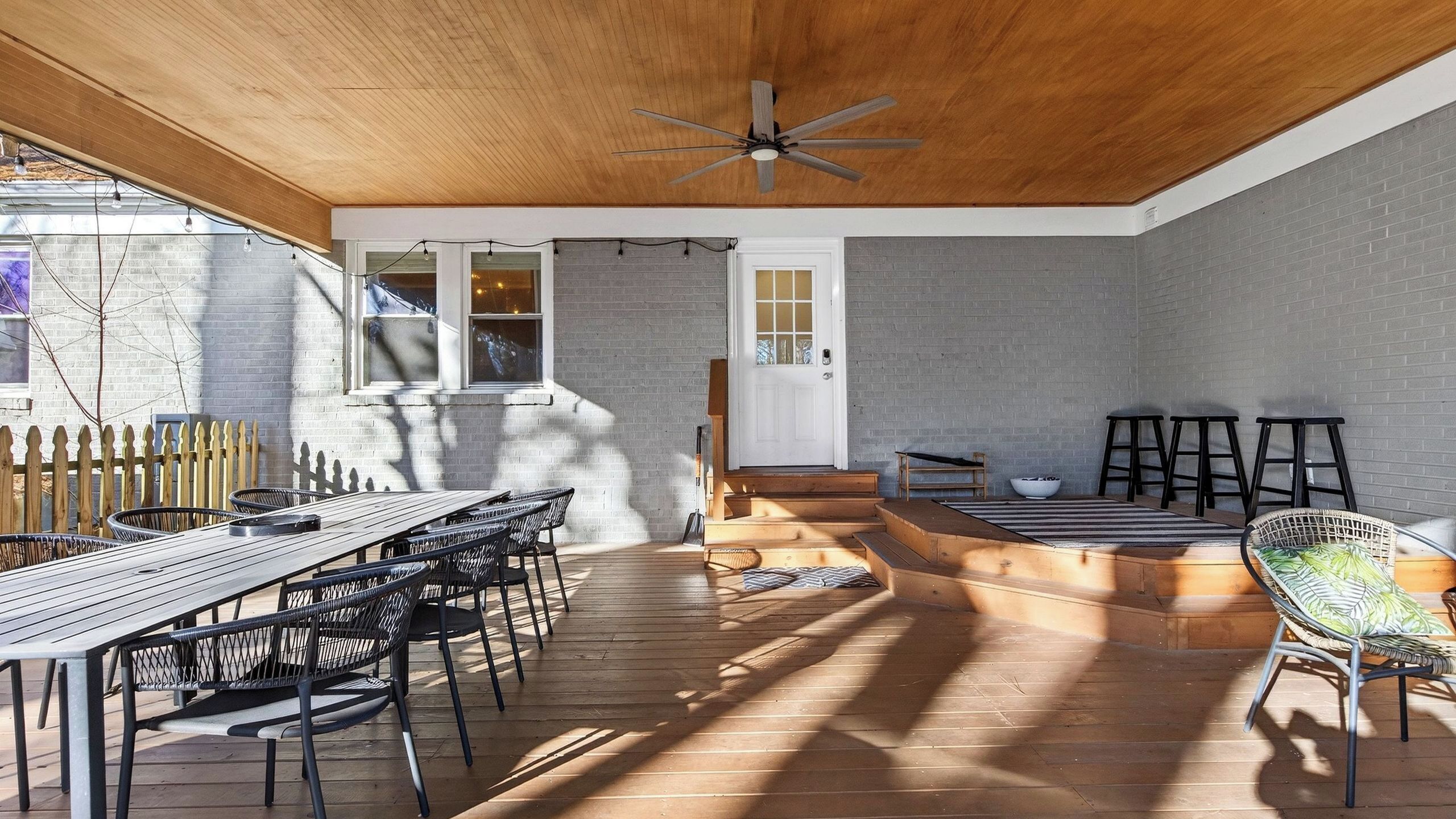 Un patio couvert avec une terrasse en bois, une table à manger avec des chaises, un coin salon avec un banc et un ventilateur de plafond.