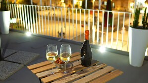 Een houten tafel met twee glazen en een fles op een balkon met uitzicht op een stadszicht 's nachts.
