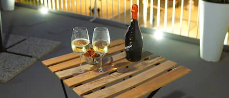 Una mesa de madera con dos vasos y una botella en un balcón con vistas a un paisaje urbano nocturno.