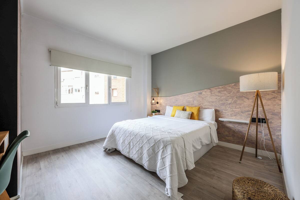 Un dormitorio con cama, lámpara de pie, ventana y suelo de madera.