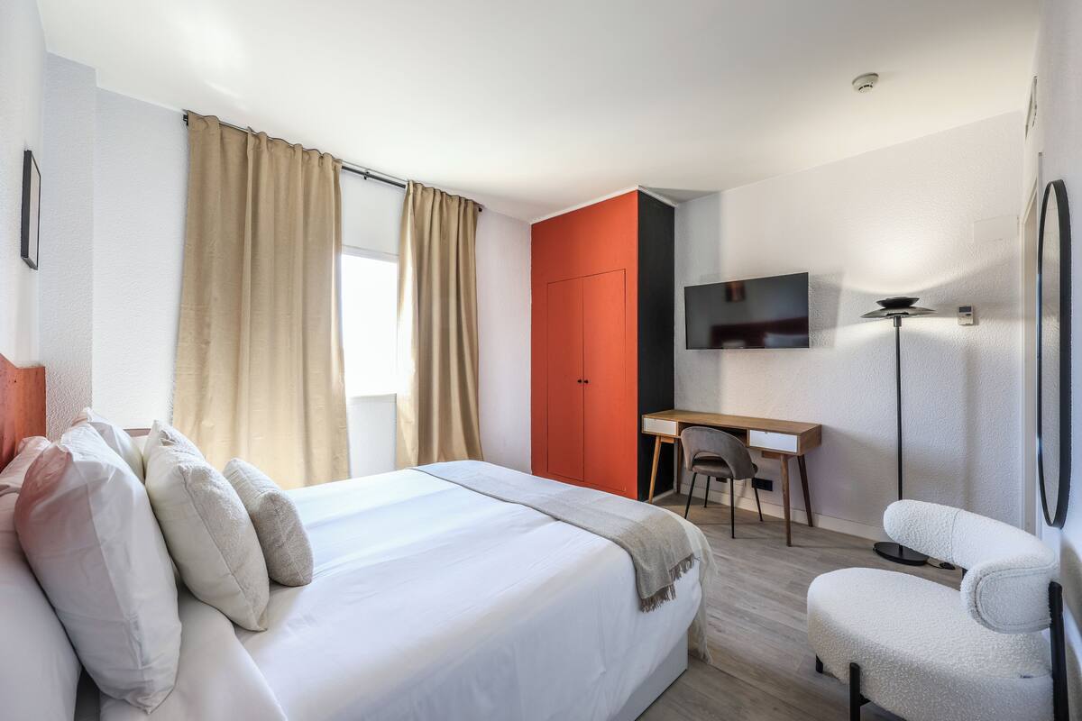 Habitación de hotel con cama, escritorio, silla, televisor y ventana con cortinas.
