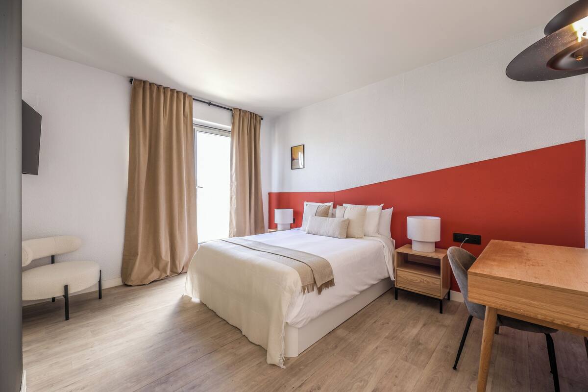 Una habitación de hotel con cama, mesitas de noche, escritorio, silla y ventana con cortinas.