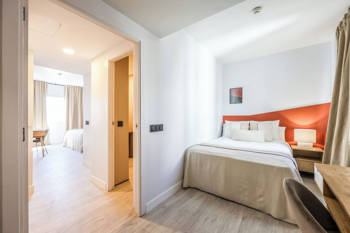 Una habitación de hotel con cama, mesita de noche, silla y vista a otra habitación.