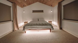 Ein Schlafzimmer mit schräg steilendem Dach, einem Bett mit Kopfteil, Nachttischlampen und einem Fenster mit Vorhängen.