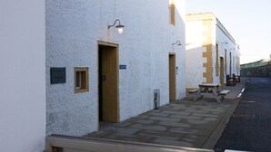 A row of white buildings with doors and windows, a paved walkway, and outdoor furniture.