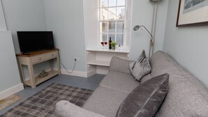 A living room with a grey sofa, a wooden TV stand, a small table with a wine bottle, and a window with a view of buildings.