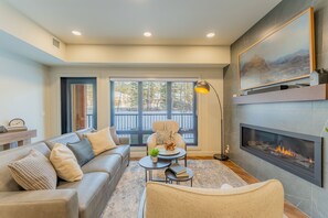 A modern living room with a fireplace, a sofa, a coffee table, and a large window. - 4BR Canmore Condo w/ Pool, Hot Tub & Gym Access (Canmore)