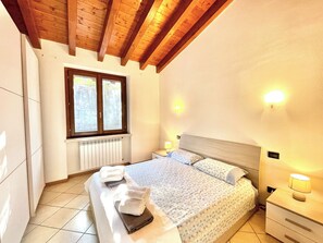 Ein Schlafzimmer mit Holzboden, einem Bett mit gemustertem Überwurf, zwei Nachttischlampen und einem Fenster mit Jalousien.