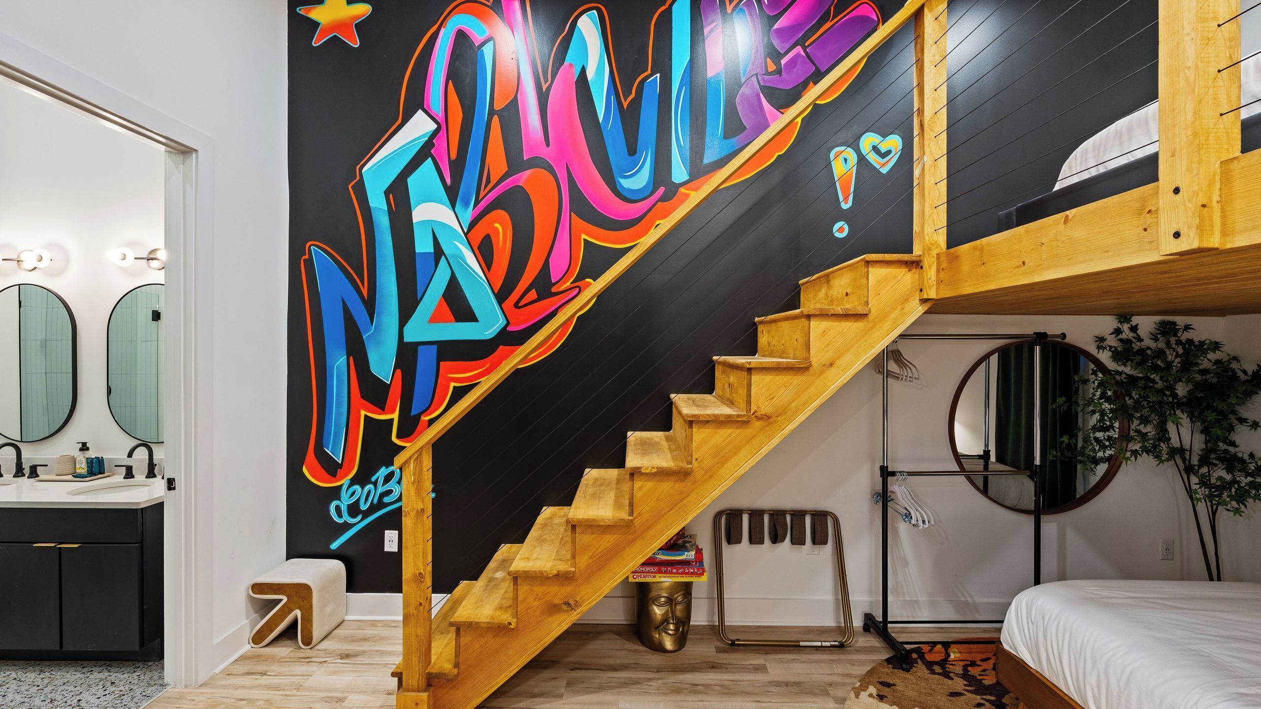 A modern interior with a wooden staircase, a black wall with colorful graffiti, a bed, and a bathroom with a mirror.