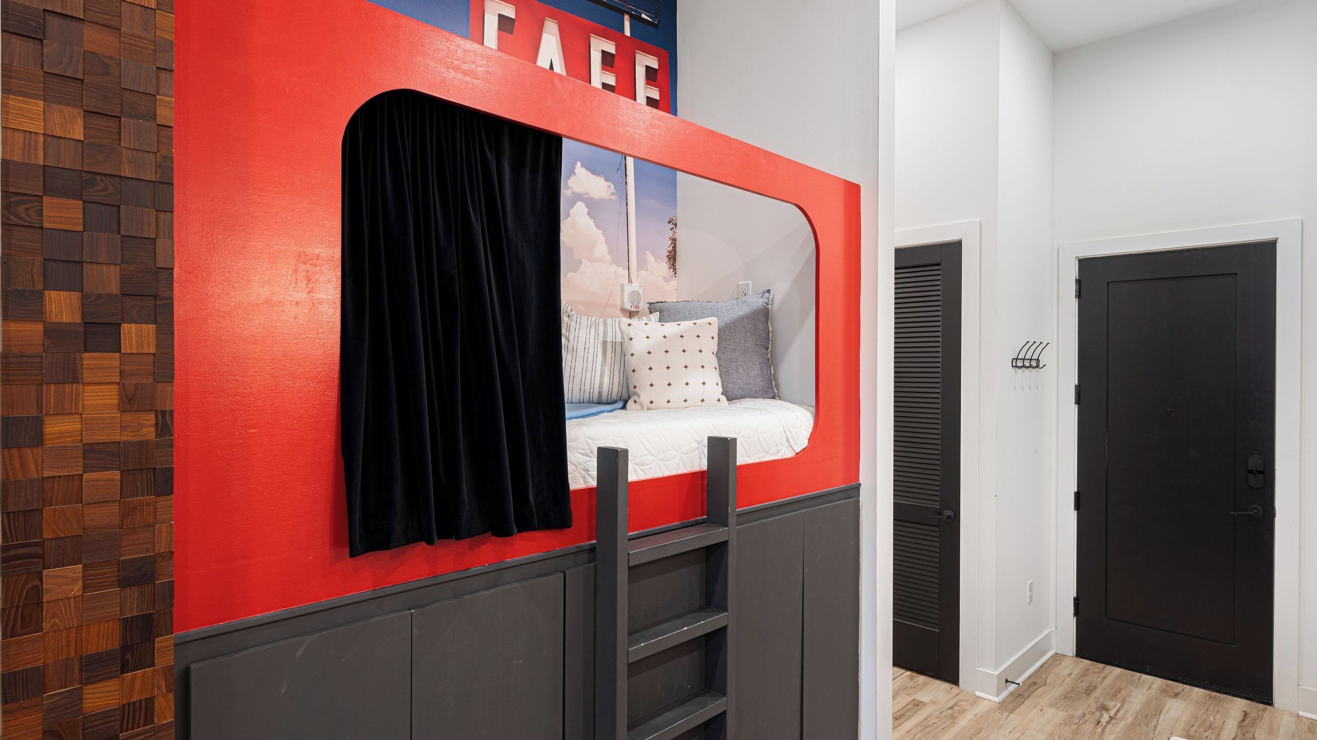 A room with a bed against a red wall, a black door, and a white wall with a window.