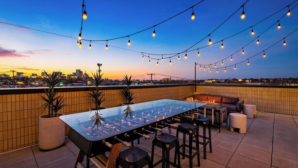 A rooftop bar with a pool table, seating, and string lights against a city skyline.