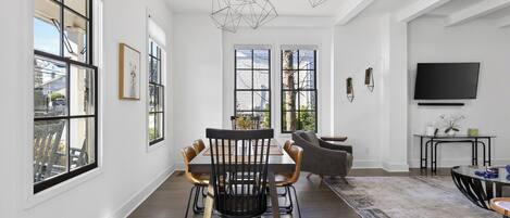 Una habitación amplia con ventanas grandes, una zona de comedor con mesa y sillas, un sofá y un televisor fijado en la pared.