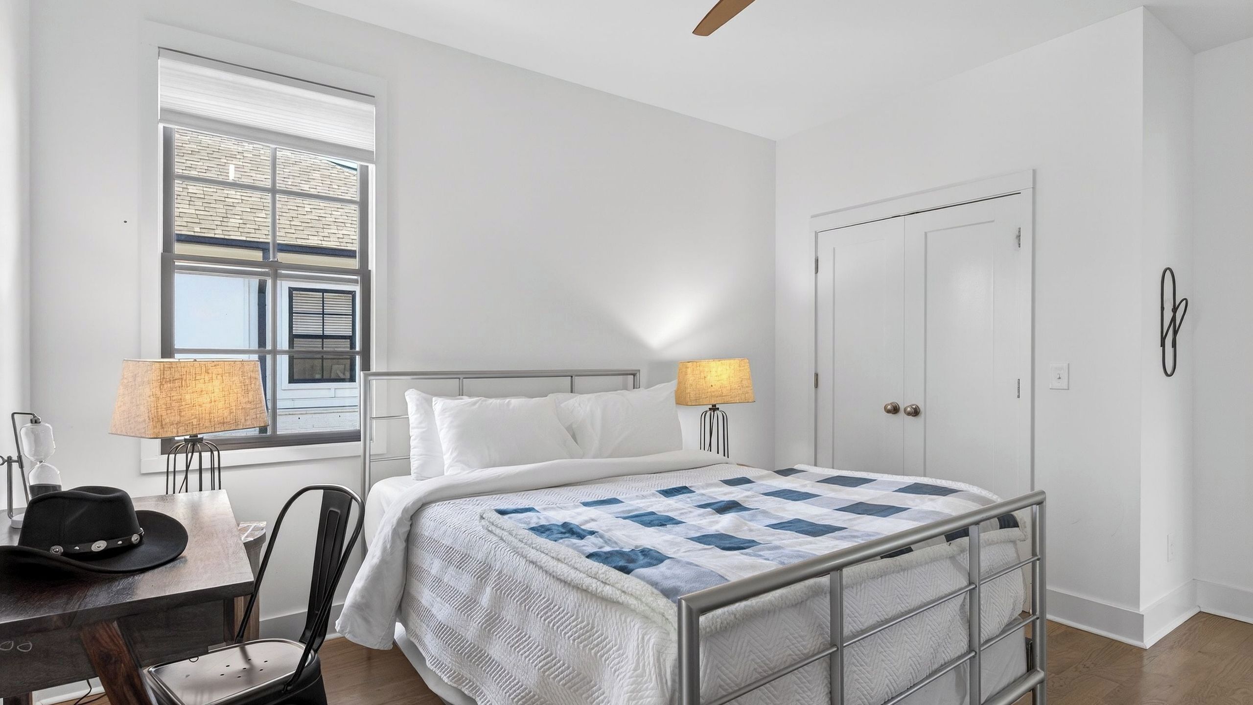 A hotel room with a bed, two bedside lamps, a chair, a desk with a hat, and a window with blinds.