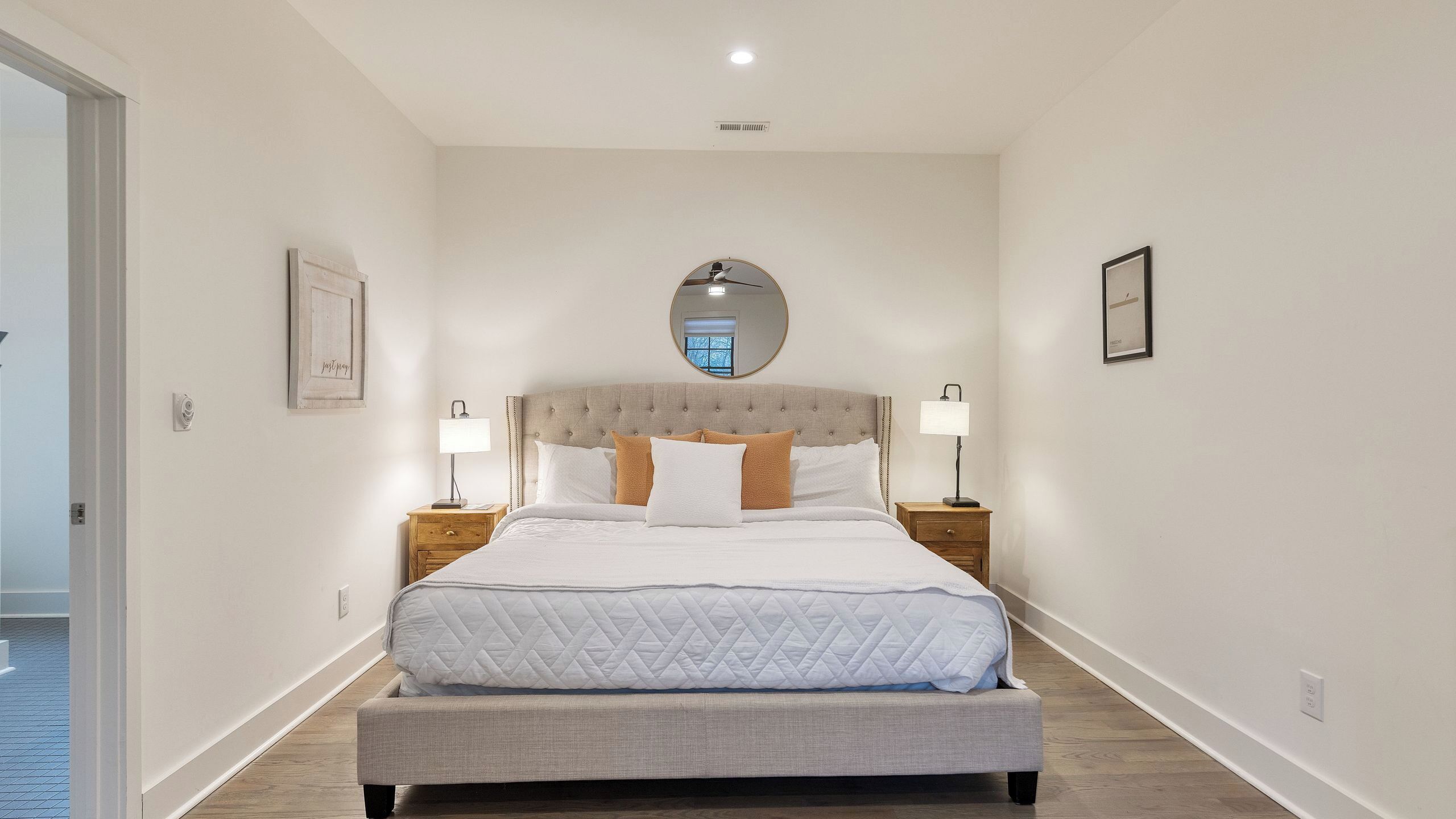 Een slaapkamer met een queensized bed, twee nachtkastjes met lampen, een ronde spiegel boven het bed en een lijst met een afbeelding aan de muur.