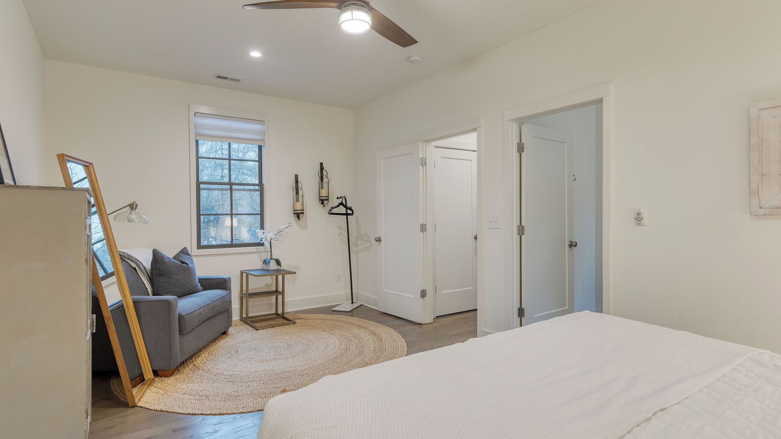 A bedroom with a bed, a sofa, a small table, a floor lamp, and a window with blinds.