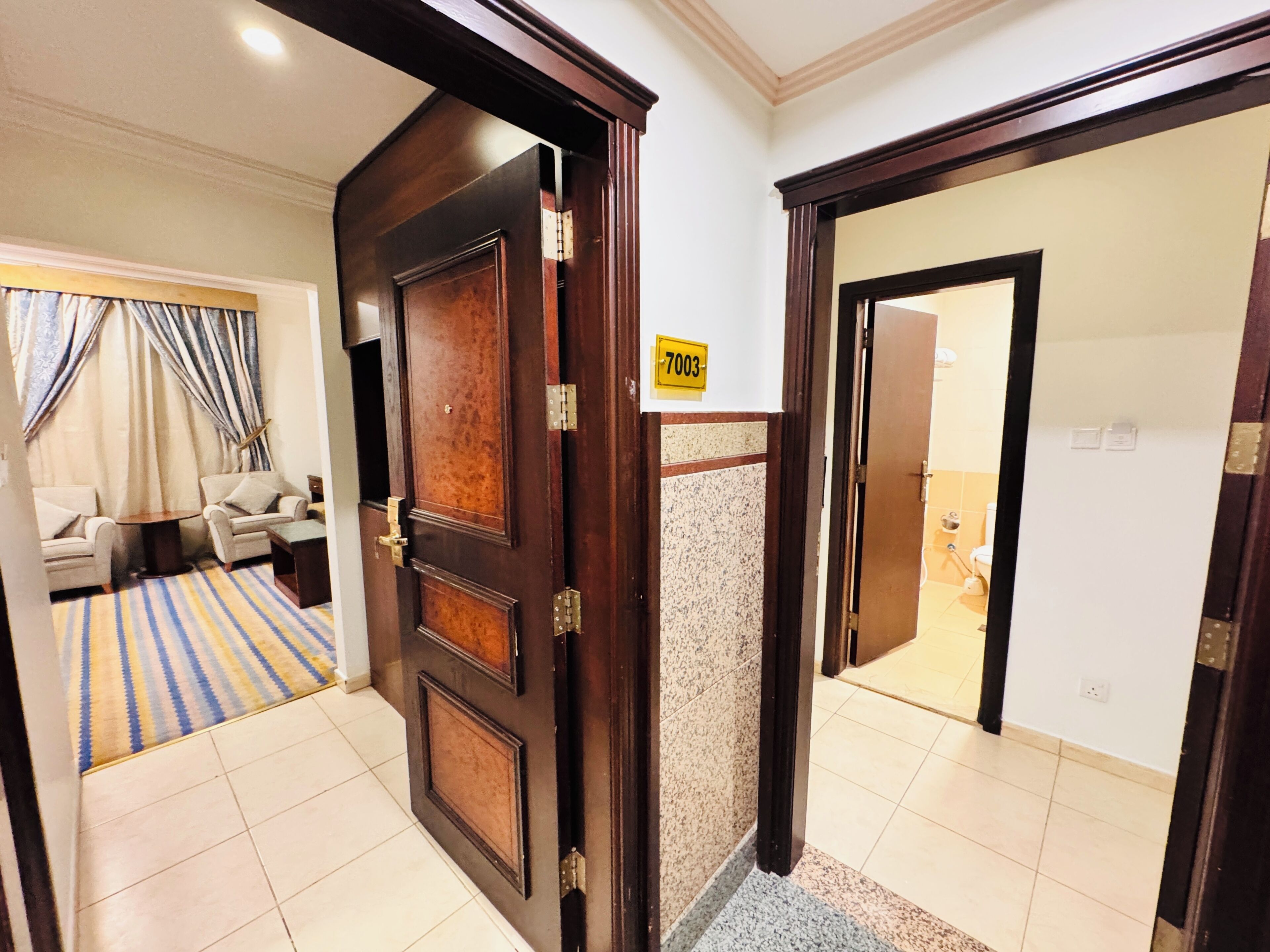 Een hotelkamer met een houten deur, een bord met het nummer 7003, een badkamer met toilet en wastafel, en een zithoek met een bank en gordijnen.
