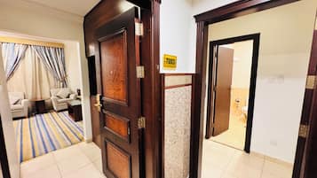Een hotelkamer met een houten deur, een bord met het nummer 7003, een badkamer met toilet en wastafel, en een zithoek met een bank en gordijnen.